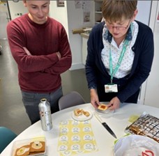 Judges Tom and Fiona from Lancaster University Library deliberate on the baking competition. On the table in front of them you can see shortbread biscuits baked to look like mini disks, a CD-Rom victoria sponge cake, a CSV file apple cake and Fiona is holding a biscuit made to look like a lantern slide.