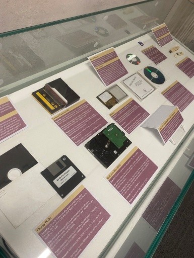 This image shows one of the cabinets from our 2025 exhibition. Inside you can see lots of obsolete hardware with descriptions such as a floppy disk, cassette tape, mini disk, CD-ROM, etc.